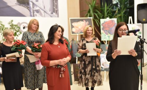 Zdjęcie przedstawia grupę kobiet podczas wernisażu. Jedna z kobiet przemawia do mikrofonu, trzymając kartki z tekstem. Pozostałe kobiety stoją obok, uśmiechając się i trzymając kwiaty oraz dyplomy. W tle widoczne są obrazy z motywami kwiatowymi oraz elementy dekoracyjne wnętrza.