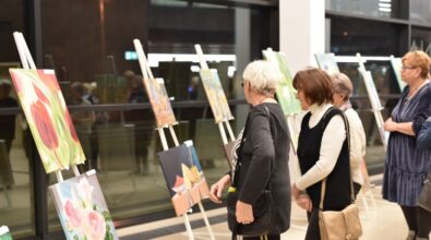 Kilka kobiet w średnim wieku ogląda obrazy ustawione na sztalugach w przestronnym wnętrzu z dużymi oknami. Na pierwszym planie widoczne są malowidła przedstawiające motywy kwiatowe oraz martwą naturę. W tle widać odbicia w szybach oraz inne osoby uczestniczące w wydarzeniu