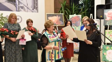 grupa kobiet odbiera wyróżnienia. Jedna z kobiet w kolorowym stroju odbiera dyplom od osoby stojącej przy mikrofonie. Pozostałe kobiety trzymają w rękach kwiaty i upominki, uśmiechając się. W tle widać obrazy o tematyce kwiatowej, a także dekoracje wnętrza, które podkreślają uroczysty charakter wydarzenia.