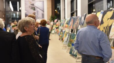 wernisaż wystawy malarskiej, odbywający się w eleganckim, przestronnym wnętrzu z dużymi oknami. Na ścianach i sztalugach rozmieszczone są różnorodne obrazy, które przyciągają uwagę zgromadzonych gości. Widzowie, w różnym wieku, przyglądają się eksponatom, niektórzy w skupieniu analizują detale dzieł sztuki. W tle widać żywą atmosferę, a światło oświetla zarówno prace, jak i osoby uczestniczące w wydarzeniu.
