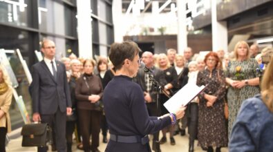 Na pierwszym planie kobieta przemawiająca do mikrofonu, trzymająca notatki w dłoniach. Za nią zgromadzona jest liczna publiczność, w tym osoby w różnym wieku, ubrane elegancko. W tle widoczne są obrazy ustawione na sztalugach oraz jasne wnętrze holu z wysokimi oknami i nowoczesnym oświetleniem.