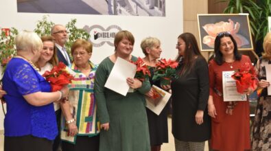 grupa osób podczas uroczystego wydarzenia. Kobiety uśmiechają się, trzymając w rękach czerwone kwiaty, dyplomy i drobne upominki. W tle widoczne są obrazy kwiatów oraz elementy dekoracyjne. Uczestnicy są elegancko ubrani, a atmosfera wydaje się radosna i uroczysta. Na ścianie za nimi znajduje się logo i elementy graficzne