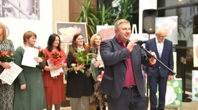 Zdjęcie przedstawia mężczyznę w garniturze przemawiającego do mikrofonu. W tle widać grupę kobiet trzymających kwiaty, dyplomy i upominki. Dekorację wnętrza stanowią obrazy kwiatów oraz rośliny