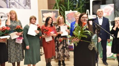 Zdjęcie przedstawia grupę kobiet i mężczyzn podczas wręczania wyróżnień. Na pierwszym planie znajduje się kobieta przy mikrofonie, trzymająca bukiet kwiatów i upominek. W tle widoczne są inne osoby z dyplomami i roślinami w doniczkach, a także obrazy przedstawiające kwiaty.