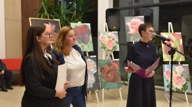 Na pierwszym planie trzy kobiety stojące przy mikrofonie – jedna z nich przemawia, trzymając w rękach różowy folder, dwie pozostałe słuchają z uśmiechem. W tle widoczne są obrazy przedstawiające motywy kwiatowe, głównie róże, ustawione na sztalugach. Dekorację wnętrza uzupełniają zielone rośliny doniczkowe. Atmosfera wydarzenia wydaje się być uroczysta i przyjazna.