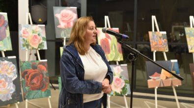 Zdjęcie przedstawia kobietę przemawiającą do mikrofonu podczas wernisażu wystawy malarskiej. Kobieta ubrana jest w białą bluzkę i jeansową kurtkę, a w tle widoczne są kolorowe obrazy przedstawiające kwiaty, martwą naturę i inne motywy, ustawione na sztalugach. Pomieszczenie ma duże, przeszklone okna, które dodają przestrzeni elegancji i światła.