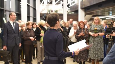 Na pierwszym planie kobieta przemawiająca do mikrofonu, trzymająca notatki w dłoniach. Za nią zgromadzona jest liczna publiczność, w tym osoby w różnym wieku, ubrane elegancko. W tle widoczne są obrazy ustawione na sztalugach oraz jasne wnętrze holu z wysokimi oknami i nowoczesnym oświetleniem.