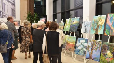 wernisaż wystawy malarskiej, odbywający się w eleganckim, przestronnym wnętrzu z dużymi oknami. Na ścianach i sztalugach rozmieszczone są różnorodne obrazy, które przyciągają uwagę zgromadzonych gości. Widzowie, w różnym wieku, przyglądają się eksponatom, niektórzy w skupieniu analizują detale dzieł sztuki. W tle widać żywą atmosferę, a światło oświetla zarówno prace, jak i osoby uczestniczące w wydarzeniu.