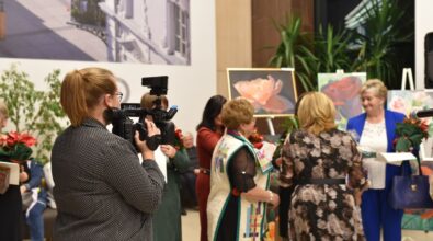 Zdjęcie przedstawia kobietę z kamerą, która rejestruje wydarzenie, podczas którego grupa kobiet odbiera wyróżnienia i upominki. Jedna z uczestniczek, ubrana w kolorową kamizelkę, rozmawia z innymi osobami. W tle widać obrazy o tematyce kwiatowej oraz dekoracje wnętrza.