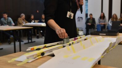 Zbliżenie na kobietę w czarnej bluzce z identyfikatorem, która trzyma mikrofon w jednej ręce, a drugą ręką umieszcza metalowe obciążenie na papierowym moście wykonanym z kolorowych, zwiniętych rurek. Most jest wzmocniony taśmą klejącą i rozpięty na drewnianym stole. W tle widoczna grupa siedzących osób, obserwujących przebieg testu.