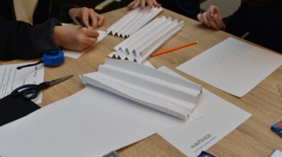 Zbliżenie na stół z materiałami do projektu podczas konkursu „GEOinżynieria”. Na blacie znajdują się złożone arkusze papieru w harmonijkę, nożyczki, taśma miernicza, ołówek i długopis, a także szkice konstrukcyjne. Uczestnicy skupiają się na pracy, rysując i przygotowując elementy projektu.