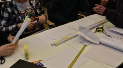 Uczniowie pracują przy stole, wykonując zadanie w trakcie konkursu „GEOinżynieria”. Na stole znajdują się zwinięte w rulon arkusze papieru, taśma klejąca, ołówek, nożyczki, zeszyt i miarka. Jeden z uczestników trzyma rulon papieru, a pozostali koncentrują się na pracy zespołowej. W tle widać innych uczestników zaangażowanych w swoje projekty.