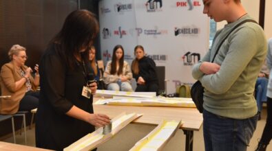 Kobieta w czarnej bluzce z identyfikatorem i mikrofonem przeprowadza test wytrzymałości papierowego mostu, umieszczając na nim obciążenie w postaci metalowych nakrętek. Obok niej stoi młody mężczyzna w zielonym swetrze, uważnie obserwując przebieg testu z założonymi rękoma. W tle siedzi grupa osób na krzesłach, w tym dwie dziewczyny w jasnych strojach, a na ścianie widoczne są banery promujące wydarzenie. Atmosfera wskazuje na edukacyjny konkurs lub warsztaty dotyczące projektowania i budowy konstrukcji.