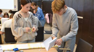 Dziewczyna i chłopak w trakcie wykonywania zadania podczas konkursu „GEOinżynieria”. Dziewczyna trzyma taśmę klejącą, przyglądając się uważnie procesowi, natomiast chłopak wycina fragment papieru. Na stole leżą przybory, takie jak kredki, nożyczki i taśma, a w tle widać innych uczestników pracujących nad swoimi projektami.