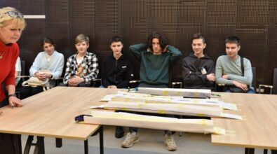 Grupa młodzieży siedzi w rzędzie na krzesłach, uważnie obserwując papierowe konstrukcje mostów umieszczone na stole przed nimi. Uczestnicy wydają się podekscytowani i skupieni, a ich różne postawy — od założonych rąk po dłonie za głową — oddają mieszankę napięcia i oczekiwania. Po lewej stronie osoba w czerwonym swetrze z pochyloną głową przygotowuje się do testu mostu.