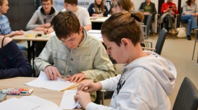 Uczestnicy konkursu „GEOinżynieria” intensywnie pracują nad zadaniem przy wspólnym stole. Dwóch chłopców w skupieniu składa kartki papieru, korzystając z linijki i innych narzędzi. W tle widać innych uczniów zaangażowanych w rozwiązywanie zadań, a także opiekunów i obserwatorów siedzących na krzesłach. Na stole znajdują się przybory szkolne, takie jak kolorowe kredki i notatki, co podkreśla edukacyjny charakter wydarzenia.