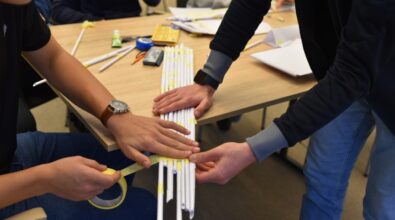 Dwóch uczestników pracuje nad wzmocnieniem mostu z papieru, przygotowując się do konkursu „GEOinżynieria”. Jeden z nich owija konstrukcję żółtą taśmą klejącą, a drugi pomaga, trzymając elementy w stabilnej pozycji. Na stole leżą przybory, takie jak linijka, klej, kolorowe kredki i miarka, potrzebne do stworzenia modelu. Widoczne są także zwinięte papierowe rurki, które stanowią podstawę budowanej konstrukcji.