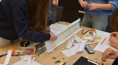 Uczestnicy konkursu „GEOinżynieria” dopracowują szczegóły papierowego mostu, używając żółtej taśmy klejącej i kolorowych ołówków. Jedna z osób pochyla się nad konstrukcją, upewniając się, że wszystkie elementy są stabilnie połączone. Na stole widać rozrzucone narzędzia, takie jak nożyczki, miarka, kredki, plastikowa ciastolina i kawałki papieru, które służą do modelowania i dekoracji. Atmosfera wskazuje na intensywną pracę zespołową i kreatywność.