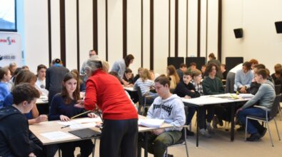 W sali pełnej młodzieży trwa konkurs drużynowy „GEOinżynieria”. Na pierwszym planie nauczycielka w czerwonym swetrze pomaga uczniom przy stole, omawiając zadanie. W tle widać kolejne grupy uczestników, skoncentrowanych na pracy zespołowej, a także opiekunów nadzorujących wydarzenie. Atmosfera w sali jest pełna zaangażowania i współpracy.