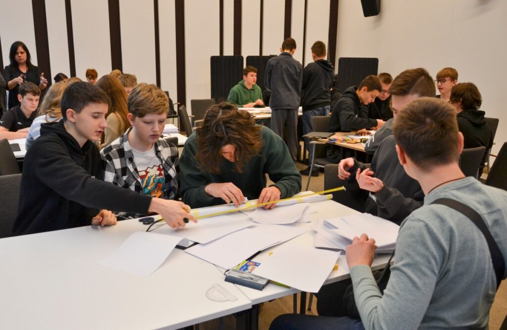 Grupa uczniów bierze udział w drużynowym konkursie matematycznym „GEOinżynieria”. Na pierwszym planie widać kilku uczestników, którzy wspólnie wykonują zadanie, używając linijki i papieru. Każdy z nich angażuje się w pracę zespołową, aby znaleźć najlepsze rozwiązanie. W tle widoczni są inni uczestnicy konkursu oraz nauczyciele i opiekunowie obserwujący wydarzenie. Sala jest wypełniona atmosferą współpracy i skupienia.