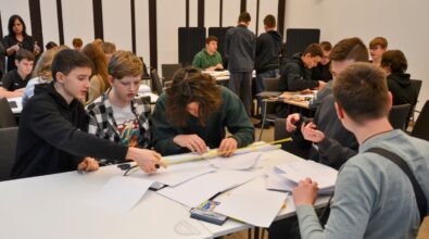 Grupa uczniów bierze udział w drużynowym konkursie matematycznym „GEOinżynieria”. Na pierwszym planie widać kilku uczestników, którzy wspólnie wykonują zadanie, używając linijki i papieru. Każdy z nich angażuje się w pracę zespołową, aby znaleźć najlepsze rozwiązanie. W tle widoczni są inni uczestnicy konkursu oraz nauczyciele i opiekunowie obserwujący wydarzenie. Sala jest wypełniona atmosferą współpracy i skupienia.