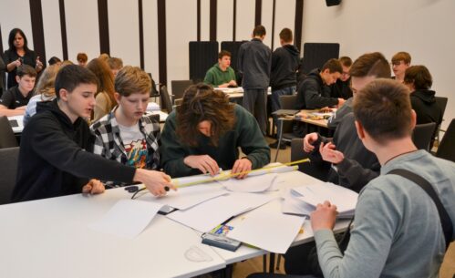 Grupa uczniów bierze udział w drużynowym konkursie matematycznym „GEOinżynieria”. Na pierwszym planie widać kilku uczestników, którzy wspólnie wykonują zadanie, używając linijki i papieru. Każdy z nich angażuje się w pracę zespołową, aby znaleźć najlepsze rozwiązanie. W tle widoczni są inni uczestnicy konkursu oraz nauczyciele i opiekunowie obserwujący wydarzenie. Sala jest wypełniona atmosferą współpracy i skupienia.