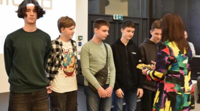 Grupa młodych osób stoi w rzędzie podczas wydarzenia. Po lewej stronie kadru znajduje się pięciu chłopców w różnorodnych ubraniach – od ciemnych bluz po koszulki z nadrukami. Na pierwszym planie z prawej strony kobieta w kolorowej marynarce przekazuje karty podarunkowe członkom grupy. W tle widoczna jest jasna ściana z napisem oraz drzwi.