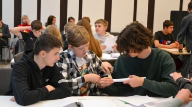 Grupa uczniów biorących udział w konkursie „GEOinżynieria” współpracuje nad rozwiązaniem zadania. Chłopcy w skupieniu używają linijki i kartki papieru, omawiając szczegóły projektu. W tle widać innych uczestników konkursu oraz opiekunów obserwujących przebieg wydarzenia. Atmosfera na sali sprzyja kreatywności i pracy zespołowej.