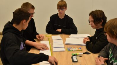 Grupa uczniów siedzących przy stole podczas pracy zespołowej na konkursie „GEOinżynieria”. Na stole znajdują się złożone arkusze papieru, taśma miernicza, kolorowe kredki, klej i inne materiały biurowe. Uczniowie omawiają swoje pomysły i przygotowują konstrukcję, pracując nad wspólnym projektem.