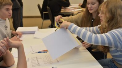 Dwie dziewczyny w trakcie kreatywnej pracy przy stole podczas konkursu „GEOinżynieria”. Jedna z nich używa taśmy klejącej do połączenia arkuszy papieru, druga uważnie obserwuje. Na stole leżą instrukcje, długopisy oraz inne przybory, w tym klej i kolorowe kredki. W tle widać innych uczestników skupionych na swoich zadaniach.