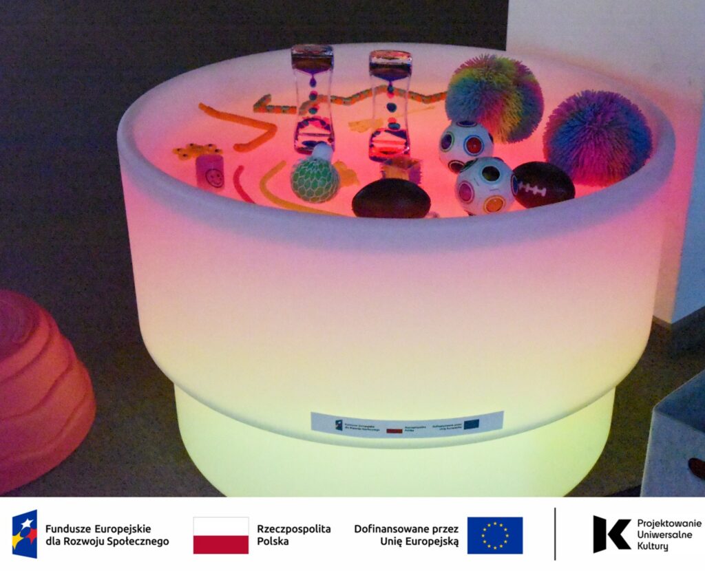 Na zdjęciu widoczny jest podświetlany stół sensoryczny stanowiący element kącika sensorycznego utworzonego w Mediatece 800-lecia w ramach działań Inkluzoteki. Na jego powierzchni znajdują się różnorodne pomoce sensoryczne o zróżnicowanych kształtach, i kolorach.