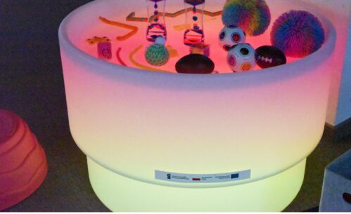 Na zdjęciu widoczny jest podświetlany stół sensoryczny stanowiący element kącika sensorycznego utworzonego w Mediatece 800-lecia w ramach działań Inkluzoteki. Na jego powierzchni znajdują się różnorodne pomoce sensoryczne o zróżnicowanych kształtach, i kolorach.