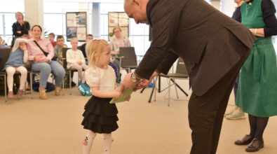 Mężczyzna w garniturze pochyla się i wręcza dziewczynce książkę lub dyplom. Dziewczynka stoi naprzeciwko, trzymając dokument obiema rękami i patrząc na mężczyznę. W tle siedzą dzieci i dorośli, obserwując sytuację. Obok stoją dwie kobiety, które uśmiechają się i przyglądają wręczeniu. Przestrzeń jest jasna, z widocznymi krzesłami ustawionymi dla uczestników wydarzenia.