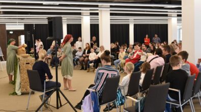 Dwie kobiety w zielonych strojach prezentują scenę teatralną w przestrzeni Mediateki. Jedna z nich stoi przy stole z koszami i rekwizytami, przygotowując elementy do przedstawienia, natomiast druga znajduje się na środku sali i zwraca się do zgromadzonej publiczności. Wokół, na ustawionych w kręgu krzesłach, siedzą dzieci oraz dorośli, którzy uważnie obserwują występ. Widzowie wypełniają znaczną część sali, a w tle widać przestronną, nowoczesną przestrzeń Mediateki z wysokim sufitem i liniowym oświetleniem.