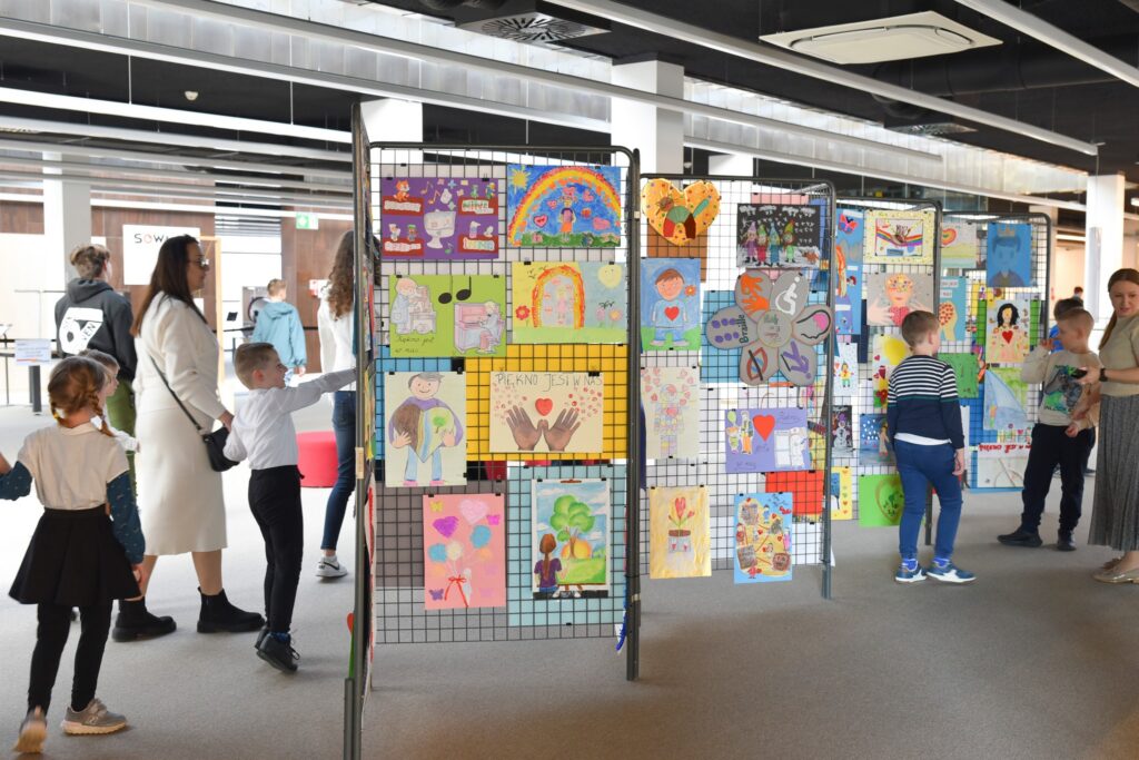 W przestrzeni Mediateki ustawione są stojaki z siatki, na których wyeksponowano kolorowe prace plastyczne wykonane przez dzieci. Rysunki przedstawiają postacie, serca, tęcze, symbole oraz różnorodne sceny, a wśród nich widoczny jest napis „Piękno jest w nas”. Wokół stojaków znajdują się dzieci i dorośli, którzy oglądają prace. Dzieci wskazują wybrane rysunki, a opiekunowie towarzyszą im podczas zwiedzania.