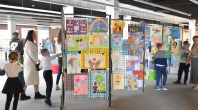 W przestrzeni Mediateki ustawione są stojaki z siatki, na których wyeksponowano kolorowe prace plastyczne wykonane przez dzieci. Rysunki przedstawiają postacie, serca, tęcze, symbole oraz różnorodne sceny, a wśród nich widoczny jest napis „Piękno jest w nas”. Wokół stojaków znajdują się dzieci i dorośli, którzy oglądają prace. Dzieci wskazują wybrane rysunki, a opiekunowie towarzyszą im podczas zwiedzania.