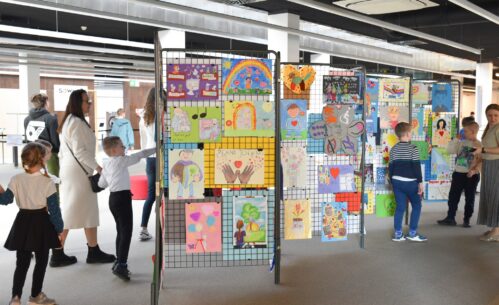 W przestrzeni Mediateki ustawione są stojaki z siatki, na których wyeksponowano kolorowe prace plastyczne wykonane przez dzieci. Rysunki przedstawiają postacie, serca, tęcze, symbole oraz różnorodne sceny, a wśród nich widoczny jest napis „Piękno jest w nas”. Wokół stojaków znajdują się dzieci i dorośli, którzy oglądają prace. Dzieci wskazują wybrane rysunki, a opiekunowie towarzyszą im podczas zwiedzania.