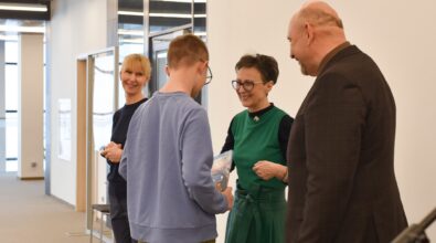 Cztery osoby stoją w przestrzeni Mediateki i rozmawiają. Młody chłopak, stojący tyłem, trzyma w rękach książkę, którą przekazuje kobiecie w zielonej sukni. Obok nich znajduje się mężczyzna w garniturze oraz druga kobieta, którzy uśmiechają się i obserwują sytuację.