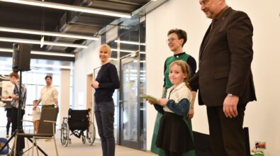 Grupa dorosłych stoi obok dziewczynki trzymającej książkę. Dziewczynka znajduje się na pierwszym planie i patrzy w stronę obserwujących. Za nią stoją dwie kobiety oraz mężczyzna w garniturze, którzy uśmiechają się i towarzyszą jej. W tle widoczna jest przestrzeń Mediateki z przeszklonymi drzwiami, oświetleniem sufitowym, krzesłem oraz wózkiem inwalidzkim ustawionym przy ścianie.