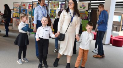 Dzieci wraz z opiekunkami spacerują między stojakami prezentującymi wystawę prac plastycznych. Kolorowe rysunki zawieszone na metalowych panelach przedstawiają m.in. postacie, serca, słońce oraz sceny z wyobraźni. Jedno z dzieci uśmiecha się i trzyma opiekunkę za rękę, podczas gdy pozostałe przyglądają się pracom lub rozmawiają ze sobą. W przestronnej sali wystawowej w tle widoczne są kolejne plansze z ilustracjami oraz osoby oglądające ekspozycję.