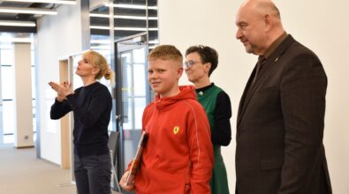 Grupa osób stoi w przestrzeni Mediateki. Na pierwszym planie chłopiec w czerwonej bluzie trzyma w ręku książkę i uśmiecha się. Obok niego znajdują się dwie kobiety oraz mężczyzna w garniturze. Jedna z kobiet wykonuje gesty dłoni, a pozostali stoją obok, zwróceni w tym samym kierunku. W tle widoczna jest nowoczesna, jasna przestrzeń z przeszklonymi ścianami i oświetleniem sufitowym.