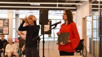 Kobieta w czerwonej bluzce prowadzi spotkanie, stojąc przy statywie z mikrofonem i trzymając w ręku teczkę z notatkami. Obok niej znajduje się tłumaczka polskiego języka migowego (PJM), która przekazuje treść wystąpienia za pomocą gestów i mimiki. W pierwszym rzędzie siedzą dzieci obserwujące wystąpienie i tłumaczenie. W tle widoczne są plansze z plakatami, duże okna oraz przestrzeń Mediateki, a z boku ustawiony jest wózek inwalidzki, podkreślający dostępność wydarzenia dla różnych uczestników.