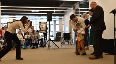W przestrzeni Mediateki odbywa się wydarzenie z udziałem dzieci i dorosłych. Na pierwszym planie kobieta pochyla się z aparatem fotograficznym, wykonując zdjęcie. Naprzeciw niej stoi mały chłopiec trzymający książkę, wspierany przez dorosłego mężczyznę. Obok znajdują się kolejne osoby, które obserwują sytuację i uśmiechają się. W tle widoczna jest grupa dzieci siedzących i przeglądających książki, a także wózek inwalidzki ustawiony przy ścianie.