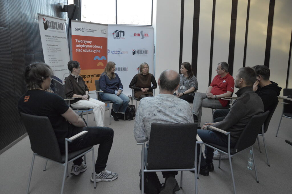Grupa kilkunastu osób siedzi na krzesłach ustawionych w kręgu i uczestniczy w spotkaniu lub dyskusji. Uczestnicy zwróceni są do siebie twarzami i prowadzą rozmowę w spokojnej, kameralnej atmosferze. W tle stoją roll-upy z logotypami Mediateki, Kina Piksel oraz Miasta Piotrków Trybunalski, a także baner z hasłem „Tworzymy międzynarodową sieć edukacyjną”. W centralnej części kręgu siedzą osoby prowadzące rozmowę, natomiast pozostali uczestnicy słuchają i włączają się do dyskusji.