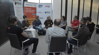 Grupa kilkunastu osób siedzi na krzesłach ustawionych w kręgu i uczestniczy w spotkaniu lub dyskusji. Uczestnicy zwróceni są do siebie twarzami i prowadzą rozmowę w spokojnej, kameralnej atmosferze. W tle stoją roll-upy z logotypami Mediateki, Kina Piksel oraz Miasta Piotrków Trybunalski, a także baner z hasłem „Tworzymy międzynarodową sieć edukacyjną”. W centralnej części kręgu siedzą osoby prowadzące rozmowę, natomiast pozostali uczestnicy słuchają i włączają się do dyskusji.