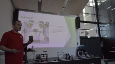 Mężczyzna w czerwonej koszulce prowadzi prezentację dotyczącą drukarki 3D. Stoi obok dużego ekranu, na którym wyświetlony jest slajd zatytułowany „Drukarka 3D” z ilustracją urządzenia i opisem procesu druku. W dłoni trzyma niewielki element wydrukowany w technologii 3D i pokazuje go uczestnikom. Na stole przed ekranem ustawione są różne modele drukarek 3D oraz przykładowe wydruki i elementy konstrukcyjne. Wnętrze sali jest wysokie i dobrze oświetlone, z dużymi oknami i panelami akustycznymi na suficie.