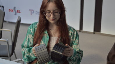 Młoda kobieta z długimi rudymi włosami i w okularach siedzi przy stole i ogląda dwa szerokie, czarne elementy przypominające ozdobne bransolety lub opaski z wypukłymi zdobieniami. Ubrana jest w zieloną koszulę z jasnym wzorem i trzyma przedmioty w dłoniach, przyglądając się ich powierzchni. W tle widoczna jest sala z krzesłami oraz ścianka z logotypami, m.in. Kina Piksel i Miasta Piotrków Trybunalski.