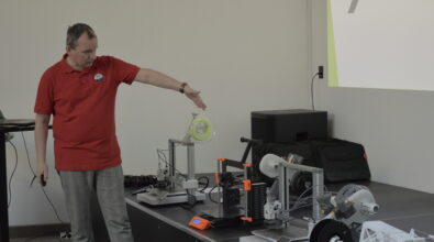 Mężczyzna w czerwonej koszulce stoi przy stole i prezentuje urządzenia techniczne ustawione obok siebie. Gestem ręki wskazuje na jeden z modeli drukarki 3D wyposażonej w szpulę z filamentem. Na stole znajdują się także inne drukarki 3D, elementy konstrukcyjne oraz akcesoria do druku. Po prawej stronie widoczny jest ekran z wyświetlaną prezentacją, a obok stoją głośnik i torby ze sprzętem.