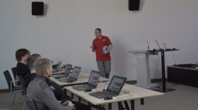 Mężczyzna w czerwonej koszulce stoi przed uczestnikami szkolenia i prowadzi prezentację. W ręku trzyma szpulę z filamentem do drukarki 3D i wyjaśnia jej zastosowanie. Przed nim przy stołach siedzą uczestnicy z otwartymi laptopami, którzy obserwują prowadzącego i słuchają wykładu. Po prawej stronie znajduje się mównica z komputerem oraz sprzętem multimedialnym, a na ścianie zamontowane są głośniki.