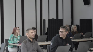 Uczestnicy szkolenia siedzą przy stołach z laptopami i słuchają prowadzącego. W pierwszym rzędzie dwaj mężczyźni rozmawiają i uśmiechają się, mając przed sobą otwarte komputery. W kolejnych rzędach siedzą inne osoby pracujące na laptopach i obserwujące prezentację. Po lewej stronie widoczna jest młoda kobieta z długimi rudymi włosami w zielonej koszuli z jasnym wzorem, która podpiera głowę dłonią i patrzy w stronę prowadzącego. W sali ustawione są stoły z komputerami, a na ścianie znajdują się panele akustyczne oraz głośniki.